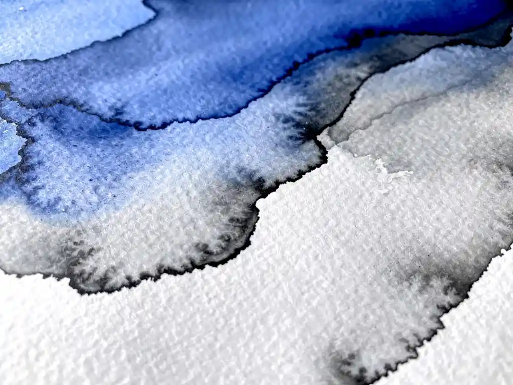 Waterverf in elektrisch blauw en houtskoolgrijs vloeit over wit papier met zichtbare vezeltextuur en natuurlijke kleurovergangen